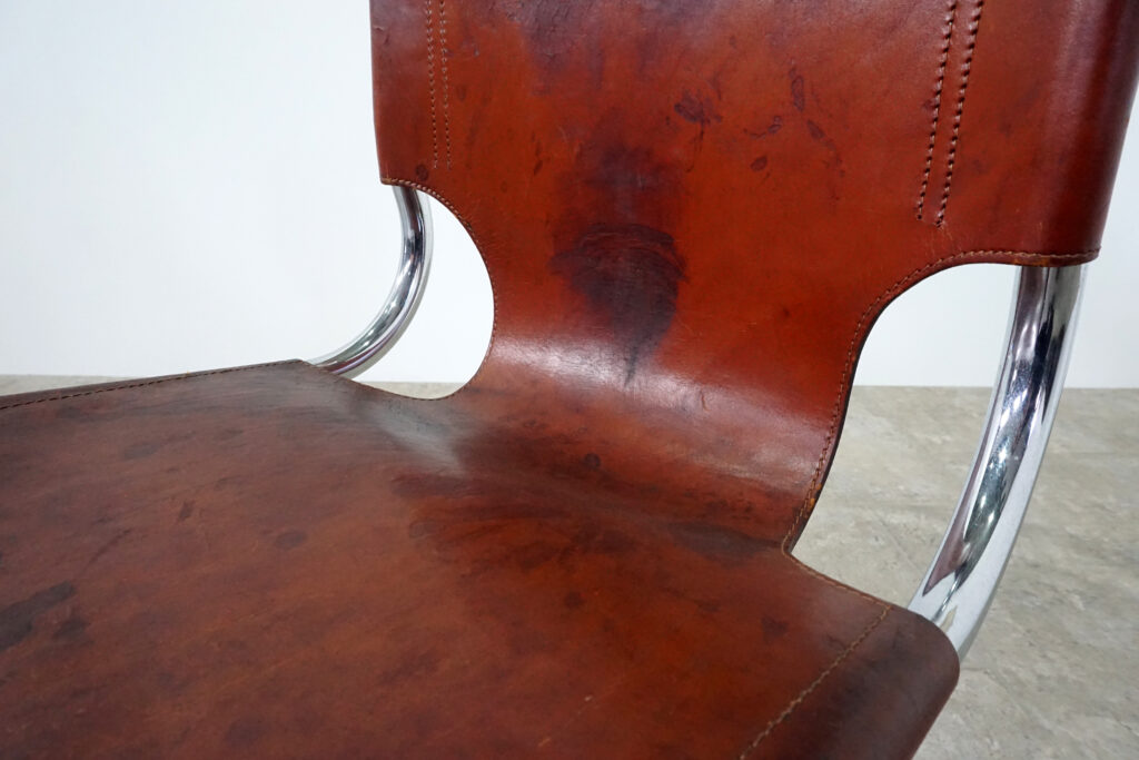 1970年代イタリア人デザイナー カルロ・バルトリ（Carlo Bartoli）によってデザインされたダイニングチェア。背もたれのアルミパーツが破損の為、より強度のあるスチール（黒クロームメッキ）を製作。深みのあるエイジングレザーと、滑らかにカーブする金属フレーム。クラシックとモダンが絶妙に交わる、スタイリッシュなダイニングチェア。座面境界