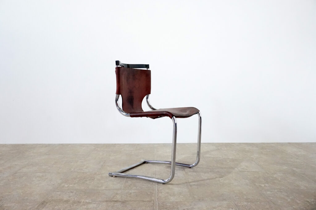 1970年代イタリア人デザイナー カルロ・バルトリ（Carlo Bartoli）によってデザインされたダイニングチェア。背もたれのアルミパーツが破損の為、より強度のあるスチール（黒クロームメッキ）を製作。深みのあるエイジングレザーと、滑らかにカーブする金属フレーム。クラシックとモダンが絶妙に交わる、スタイリッシュなダイニングチェア。右斜め横