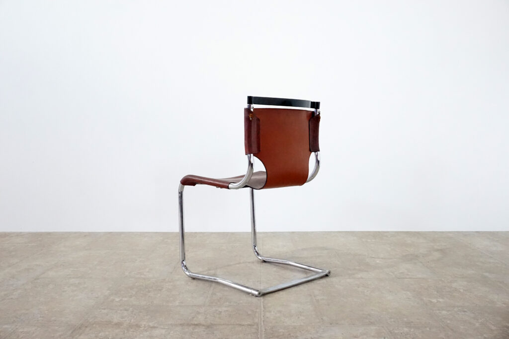 1970年代イタリア人デザイナー カルロ・バルトリ（Carlo Bartoli）によってデザインされたダイニングチェア。背もたれのアルミパーツが破損の為、より強度のあるスチール（黒クロームメッキ）を製作。深みのあるエイジングレザーと、滑らかにカーブする金属フレーム。クラシックとモダンが絶妙に交わる、スタイリッシュなダイニングチェア。左斜め後ろ