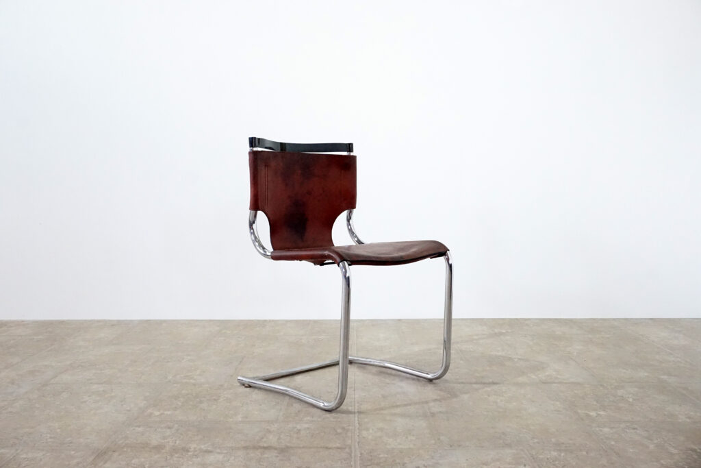 1970年代イタリア人デザイナー カルロ・バルトリ（Carlo Bartoli）によってデザインされたダイニングチェア。背もたれのアルミパーツが破損の為、より強度のあるスチール（黒クロームメッキ）を製作。深みのあるエイジングレザーと、滑らかにカーブする金属フレーム。クラシックとモダンが絶妙に交わる、スタイリッシュなダイニングチェア。左斜め前