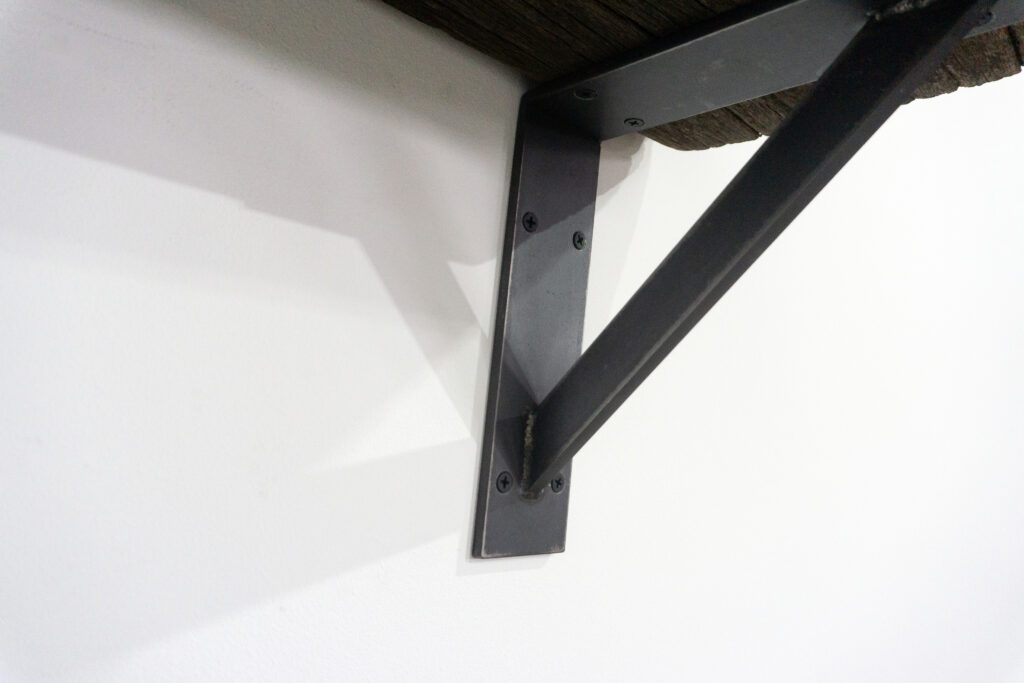 古材の梁から切り出した厚みのある木塊を、そのまま棚として再構成したウォールシェルフ。時間を刻んだ木材の質感と重量感を活かし、壁面から静かに突き出すような存在感をつくります。棚受けには、鉄の黒皮材を用いたオリジナルブラケットを新規制作。素材の表情を残した黒皮鉄を黒塗装で仕上げ、古材の力強さと調和するシンプルな構造としています。古材が持つ個体差や痕跡をそのまま活かしながら、現代の空間に静かな緊張感を与えるプロダクトです。脚横