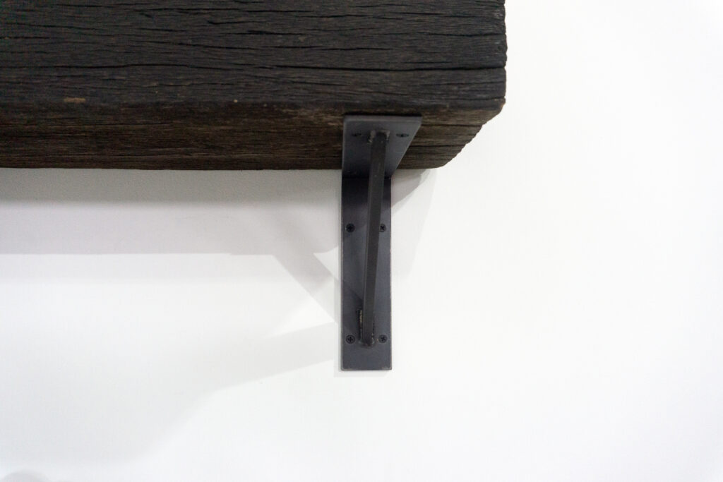 古材の梁から切り出した厚みのある木塊を、そのまま棚として再構成したウォールシェルフ。時間を刻んだ木材の質感と重量感を活かし、壁面から静かに突き出すような存在感をつくります。棚受けには、鉄の黒皮材を用いたオリジナルブラケットを新規制作。素材の表情を残した黒皮鉄を黒塗装で仕上げ、古材の力強さと調和するシンプルな構造としています。古材が持つ個体差や痕跡をそのまま活かしながら、現代の空間に静かな緊張感を与えるプロダクトです。脚正面