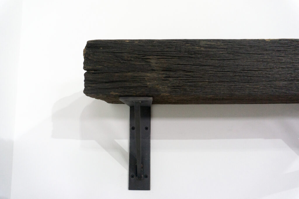 古材の梁から切り出した厚みのある木塊を、そのまま棚として再構成したウォールシェルフ。時間を刻んだ木材の質感と重量感を活かし、壁面から静かに突き出すような存在感をつくります。棚受けには、鉄の黒皮材を用いたオリジナルブラケットを新規制作。素材の表情を残した黒皮鉄を黒塗装で仕上げ、古材の力強さと調和するシンプルな構造としています。古材が持つ個体差や痕跡をそのまま活かしながら、現代の空間に静かな緊張感を与えるプロダクトです。左
